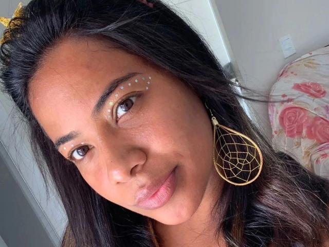 Thais Nunes, de aspecto atractivo, con ojos marrones, cabello oscuro, aretes dorados y brillo en las cejas, sonriendo ligeramente. Sus glúteos no son visibles. Lleva una diadema dorada..