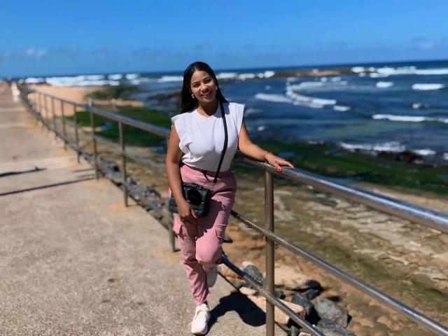 Thais Nunes – attraktiv mit langen, schwarzen Haaren, einem weißen Top ohne Ärmel, rosa Hosen, weißen Sneakers, einer schwarzen Kamera, lehnt an einem Metallgeländer, lächelt zum Meer mit den Wellen..