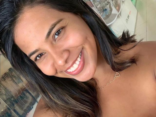 ThaisNunes-hot has dark brown hair, tan skin, and a cute smile showing white teeth. She's wearing a heart-shaped necklace, and her bare shoulders are visible.