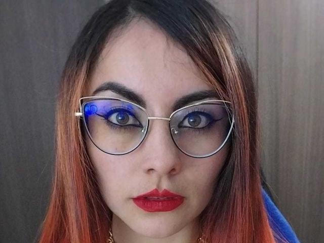 Sofia69Hoty with blue-tinted glasses, red lipstick, and orange-brown hair, looking straight at the camera, showing her gray eyes and a small nose ring on her right nostril.