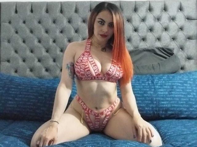 Sofia69Hoty with red and pink patterned bikini, showing her tits and pussy, sitting on blue bed with gray tufted headboard, long orange hair, tattoo on left arm.