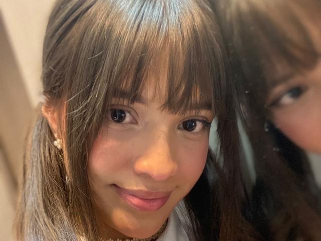 MelodySwet's face, brown hair with bangs, small nose, cute smile, brown eyes, light skin, wearing small pearl earrings, reflected in a mirror, slight blush on cheeks.