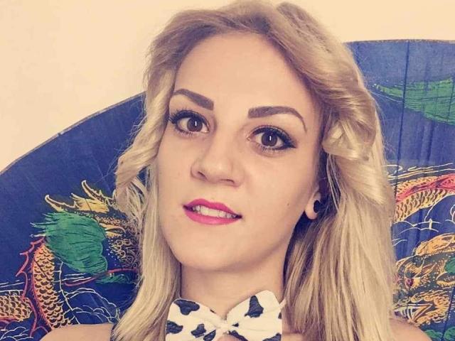 SarahFontain has blonde wavy hair, dark eyebrows, pink lipstick, and is wearing a white polka dot bowtie, with a colorful fan in the background.