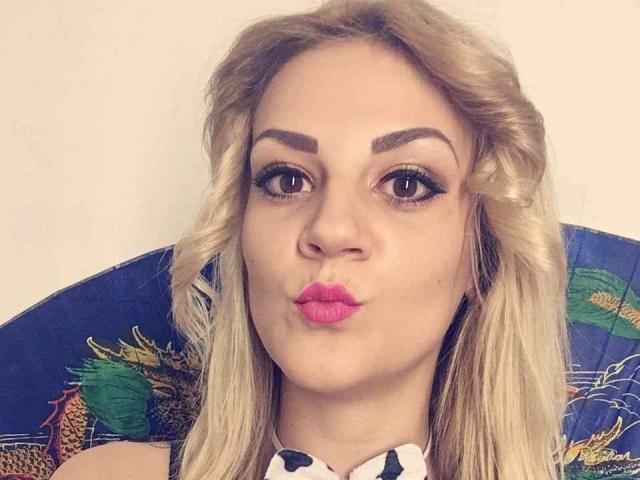 SarahFontain has blonde hair, thick eyebrows, and pink lipstick, puckering her lips in a kissy face. She's wearing a colorful, patterned top, and has dark, dramatic eye makeup.