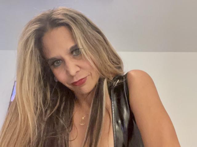 Icequren69 with long blonde and brown hair, wearing a black leather top, showing a slight smile with red lipstick, and a gold necklace, looking at the camera.