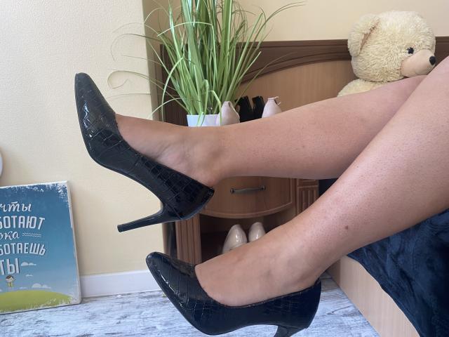 Mirellias Beine in schwarzen, glänzenden High Heels mit Krokodilmuster, die an den Knöcheln gekreuzt sind, zeigen glatte, gebräunte Haut. Im Hintergrund ein beiger Teddybär und eine grüne Pflanze..
