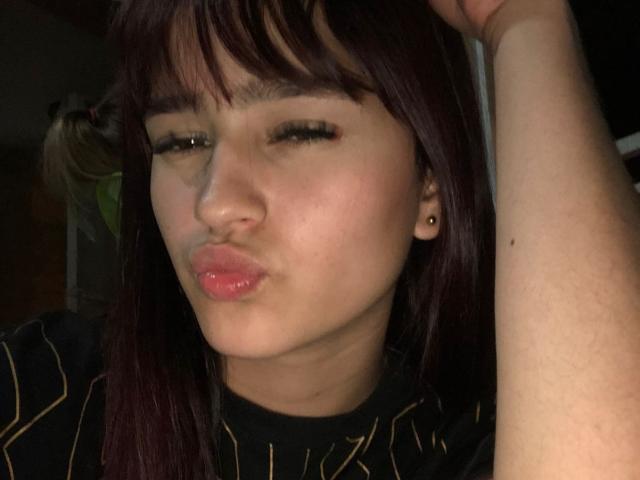 MelodyMansson has puffy, glossy lips, half-closed eyes with long lashes, and dark brown hair with bangs. She's wearing a black striped shirt, right arm raised.