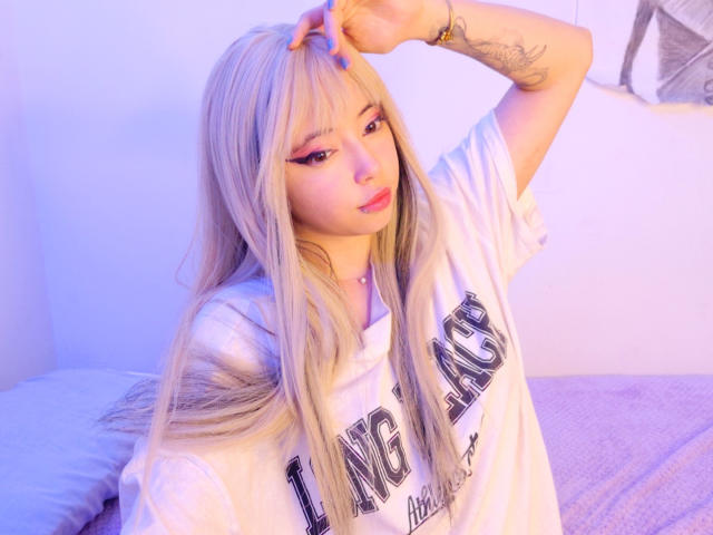 Arieyaara with long blonde hair, pink eyeliner, and blue nails, wearing a white "L.A. Angels" shirt, touching her head, showing a tattoo on her arm, in soft purple light.