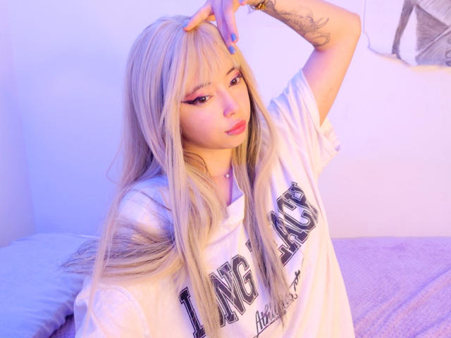 Arieyaara, con lunghi capelli biondi, indossa una felpa bianca con la scritta "UCLA", ha lo smalto di unghie blu, l'eyeliner e un tatuaggio sul braccio destro, e tocca la testa con una mano..