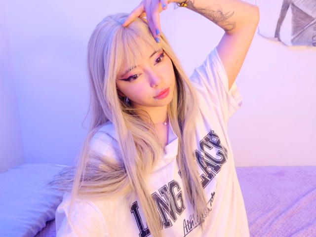 Arieyaara, con cabello rubio largo, vestida con una sudadera blanca, uñas azules y un tatuaje en el brazo, tocándose la frente, brillando con una luz púrpura-azul, sentada en una cama..