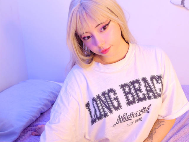 Arieyaara with blonde hair, pink eyeliner, and white Long Beach shirt, sitting on a bed, looking up with a slight smile, showing a tattoo on her right arm.