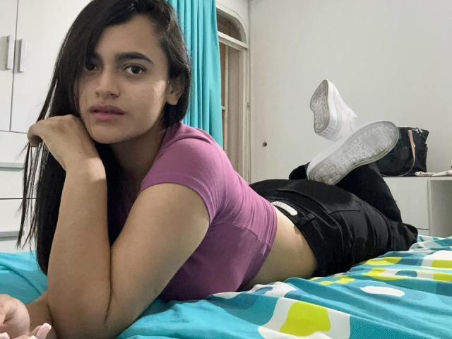 VictoriaRs, lying on a bed, wears a pink top and black shorts, showing her ass, with white sneakers and a turquoise curtain in the background.