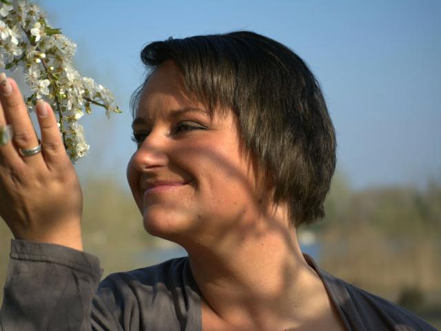 LadySun87- aantrekkelijk, met kort zwart haar, lachend, een witte bloem vasthoudend, een grijze top dragend, zonlicht op haar gezicht, een blauwe lucht op de achtergrond. Ze heeft een ring om haar linkerhand..