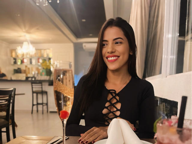 Aymens has long dark hair, red lipstick, and is smiling with white teeth. She's wearing a black lace-up top, showing her tits. She's at a restaurant, holding a napkin.