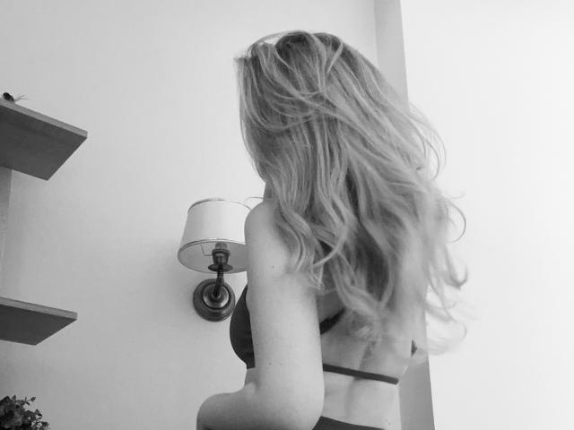 SugarrX with long, wavy blonde hair, wearing a black bra, standing with her back to the camera, showing her ass and a small, visible tattoo on her left side.