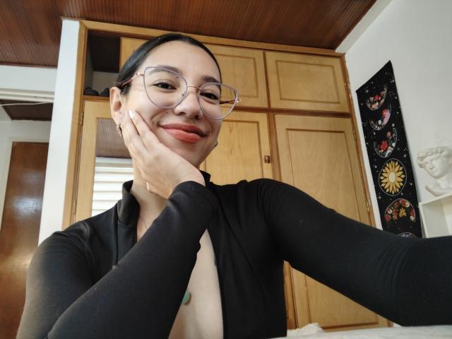 Mllevv with glasses, black top, and hand on cheek, smiling, showing a bit of her tits, in a kitchen. Wood cabinets and ceiling. She's cute and confident.