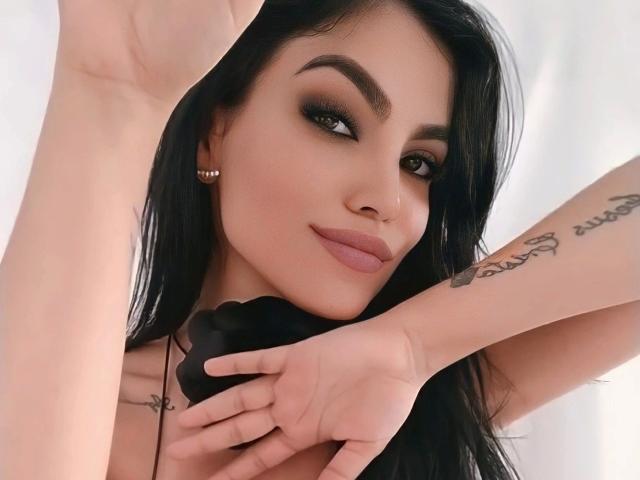 AriannaParadise-hot with black hair, green eyes, and a tattoo on her right arm, holding a black object, smiling with pink lips, and wearing gold earrings.