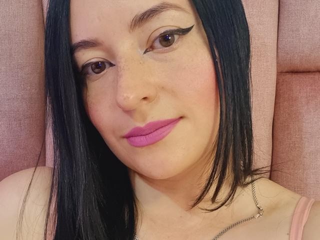 VaaleryStark a la peau pâle, les cheveux noirs et lisses, du rouge à lèvres rose, un trait d'eye-liner ailé, et un léger sourire. Elle est assise devant un fond rose texturé. Elle porte un collier fin en argent..