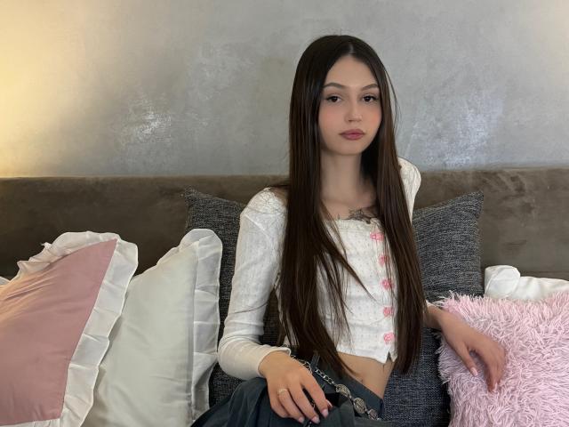 HeyWednesday, con cabello castaño largo, una camiseta blanca de botones y pantalones vaqueros azules, está sentada en un sofá gris con cojines rosa y blancos. Sus pechos son pequeños, y no se ve su trasero..