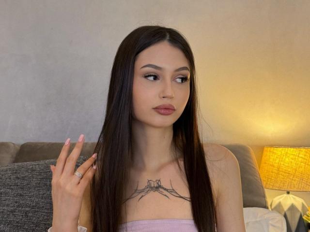 HeyWednesday, con cabello largo y oscuro, lápiz labial rosa y un tatuaje debajo de los senos, levantando un dedo, tiene uñas rosas y un anillo, sentada cerca de una lámpara amarilla..