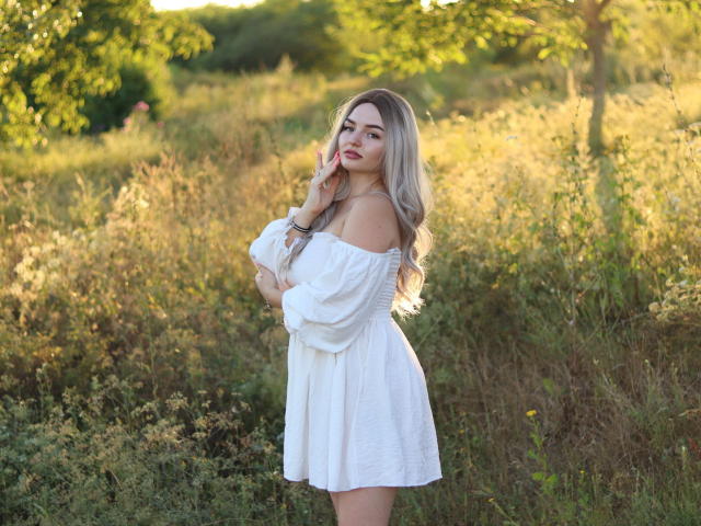 NicolPots with long blonde hair, white off-shoulder dress, standing in a sunlit field, touching her chin, showing her ass and tits, looking at the camera with a smile.