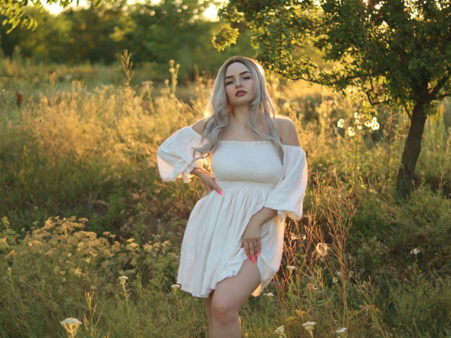 NicolPots with long blonde hair, white off-shoulder dress, pink nails, standing in sunlit field, hand on hip, slightly lifting dress, showing her bare thigh and cute ass.