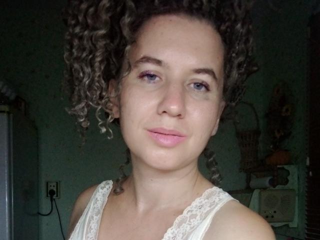 SelenaXCute with curly brown hair, light skin, and pink lipstick, wearing a white lace top, smiling softly at the camera. Her blue eyes and relaxed expression are visible.
