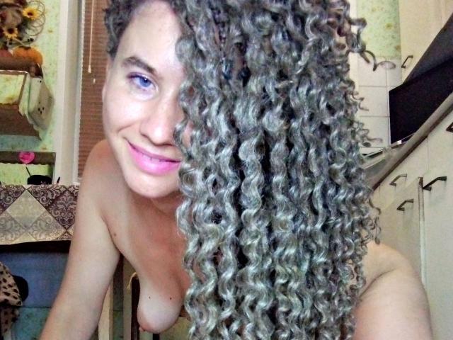 SelenaXCute with curly gray hair, smiling, topless, showing small tits, blue eyes, and pink lips, leaning forward in a kitchen.