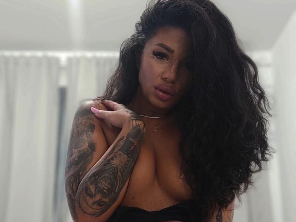 JessePureLove with long, wavy black hair, tattoos on her right arm, large bare boobs, and a serious expression, standing in a softly lit room.