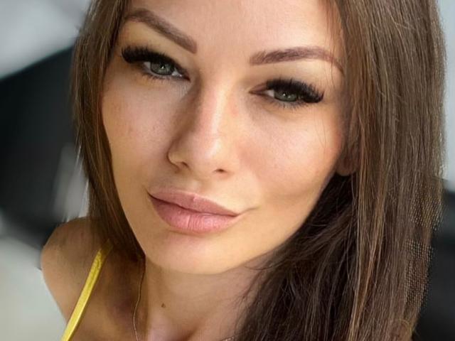 AliceMarr has light brown, straight hair, green eyes, and thick, long lashes. She's wearing a yellow top with a subtle smile, and her tits are slightly visible.