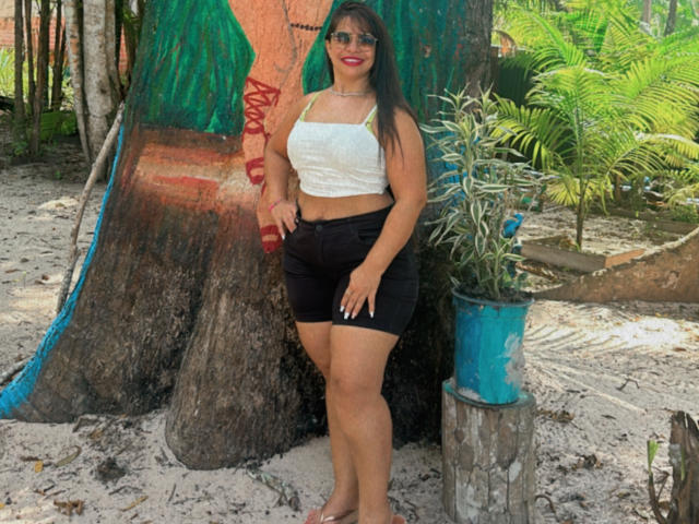 Elaini with big ass, small white tank top, black shorts, brown hair, red lipstick, standing barefoot by tree, blue pot with plant, smiling, wearing sunglasses.