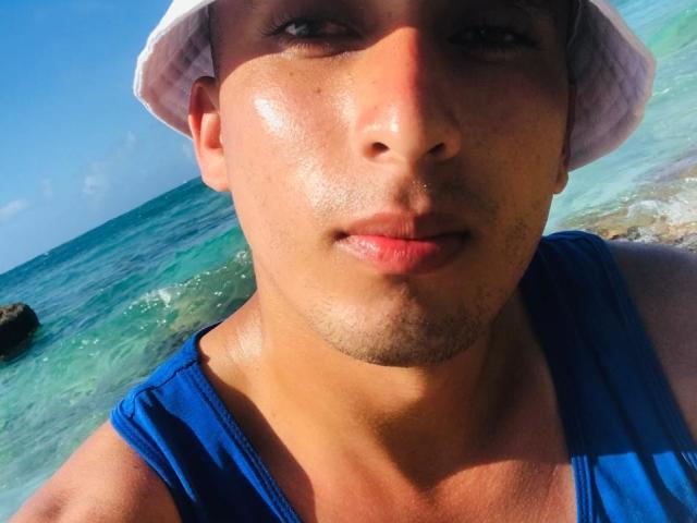 ChristoferMegia with a shiny, tan face, wearing a white bucket hat and blue tank top, showing a slight mustache, and wet lips in a beach selfie.