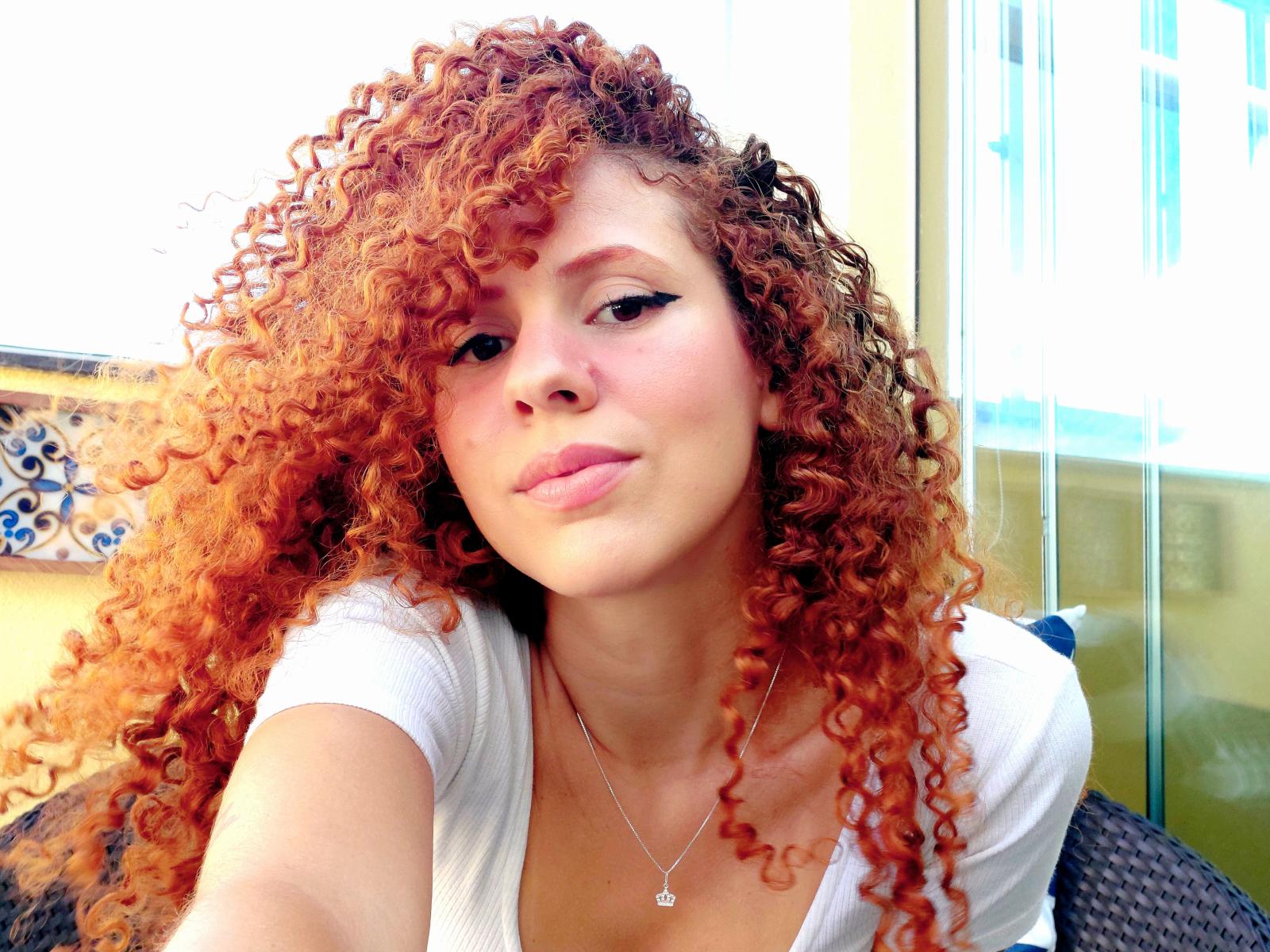 Pavani, con cabello rizado de color naranja-rojizo, una camisa blanca, labios fruncidos y un collar pequeño, está tomando una selfie, mostrando su busto y su cara linda. Luz natural brillante..