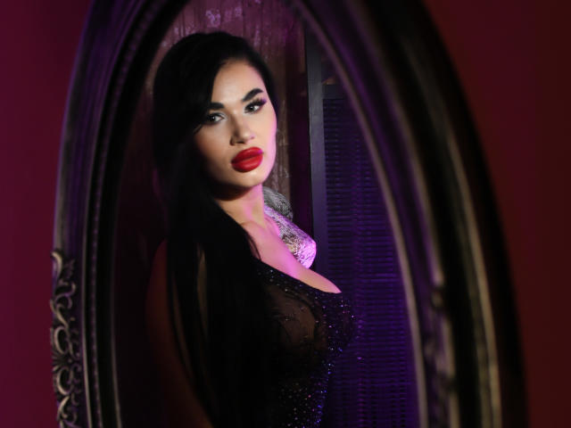LexiGlow ha un seno prosperoso, capelli scuri e rossetto rosso. Indossa un abito nero trasparente con paillettes e appare sexy riflessa in uno specchio con illuminazione viola..