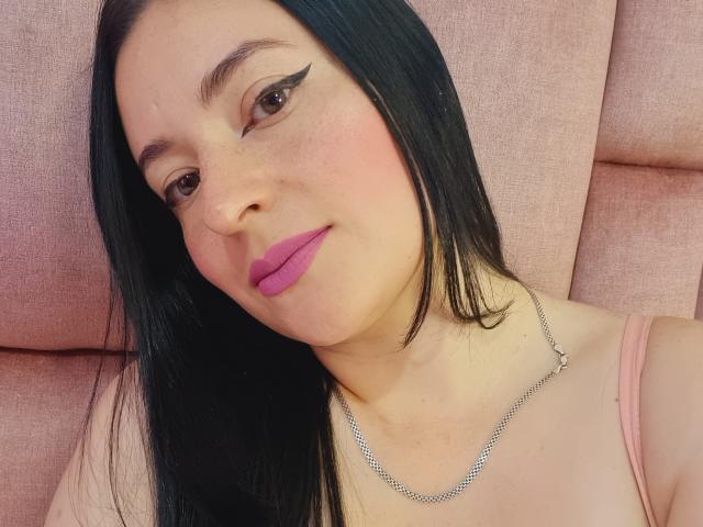 VaaleryStark has black hair, pink lipstick, and winged eyeliner, with a silver necklace, and is wearing a pink top, sitting against a pink cushioned background.