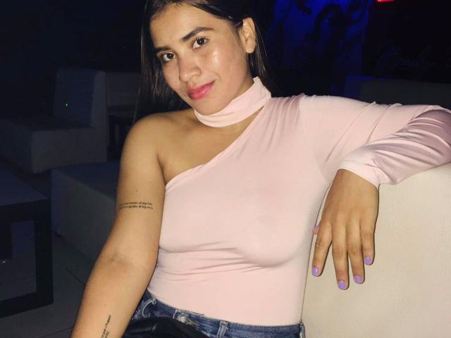 BellaBickers with medium tits, wearing a pink one-shoulder top, blue jeans, and purple nails, leaning on a white wall, smiling, dark hair, tattoo on right arm.