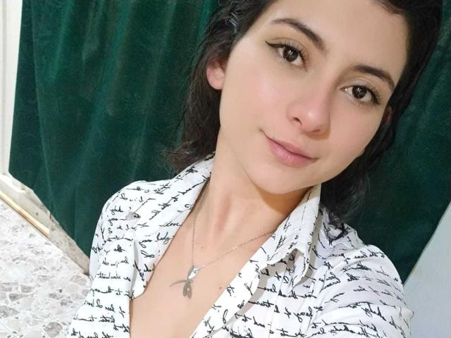 JeneKurz with small tits, wearing a white shirt with black script, black eyeliner, necklace with small pendant, smiling, dark hair, green curtain, white wall, and lace pattern.