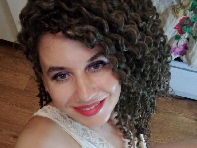 SelenaXCute with curly brown hair, purple eyeliner, red lipstick, smiling, white lace top, wooden floor, and floral-patterned bed. Boobs are not visible. Pussy is not visible.