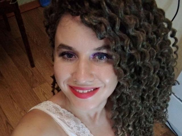 SelenaXCute has curly brown hair, purple eyeshadow, red lipstick, and a white lace top, smiling at the camera with blue eyes, wooden floor in the background.