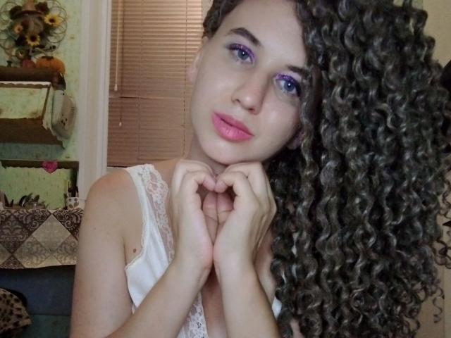 SelenaXCute with curly brown hair, purple eyeliner, pink lipstick, white lace top, hands near chin, looking at camera, blue eyes, slight smile, soft light, indoor setting.