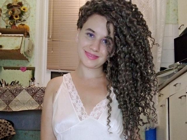 SelenaXCute with long curly brown hair, purple eyes, and pink lipstick, wearing a white lace-trimmed top, smiling in a kitchen. Boobs visible under the top.