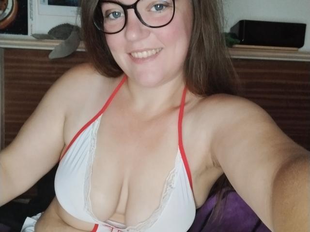 FrancaisePureSouche with glasses, smiling, showing her tits in a white, red-trimmed bikini top, brown hair, fair skin, and a cute, chubby face.