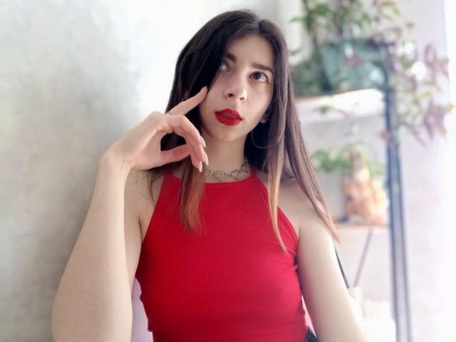 WollyMolly com cabelos castanhos longos, batom vermelho, blusa vermelha sem mangas, brincos de argola, tocando o nariz com o dedo direito, pele clara, maquiagem discreta e um colar..