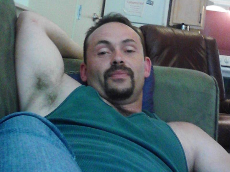 A photo of SteelRodNYou, a chubby man with a green tank top, relaxed on a couch, arm behind head, showing armpit hair, slight mustache, and a lazy smile.