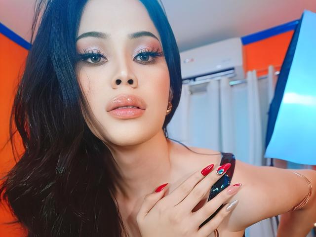 MariaWildCummer with glossy pink lips, dark wavy hair, and red nails, holding a phone, wearing a strapless top, showing off her tits. Orange and blue background.