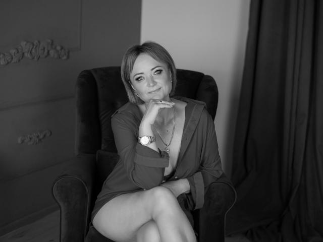 SnezhanaRells sitting in a chair, wearing a low-cut shirt, exposing her tits, with one leg crossed, resting her chin on her hand, smiling. Black-and-white photo.