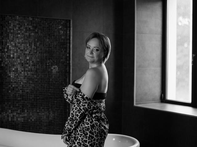 SnezhanaRells with short hair, smiling, holding a leopard print robe, exposing her black bra-covered tits, standing in a dark bathroom, near a window. Black and white photo.
