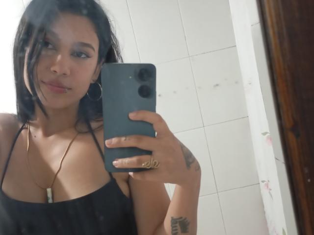KataleyaMansson with black hair, small tits, hoop earrings, gold necklace, and tattoo on right arm, taking a mirror selfie with a blue phone, smiling slightly.