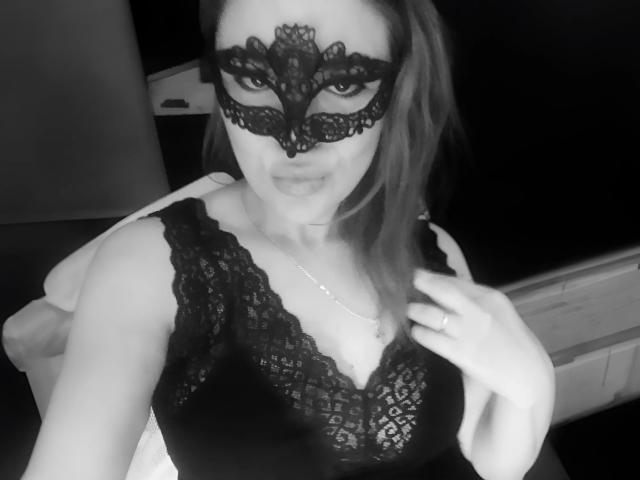 SofiaBells wearing a black lace mask, black lace top, and a silver necklace, with a hand in her hair, showing off her tits, in a black-and-white selfie.