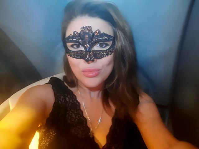 SofiaBells with a black lace mask, pouty lips, and wavy brown hair, wearing a black lace top, holding her tits, glowing yellow light in the background.